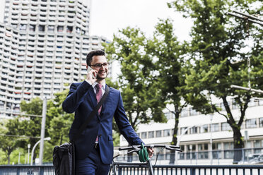Businessman with bicycle walking in the city, using smart phone - UUF007995