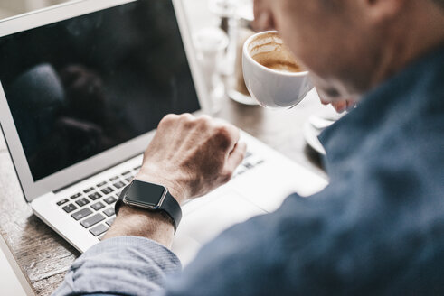 Älterer Mann trinkt Kaffee, während er am Laptop arbeitet und auf seine Smartwatch schaut - KNSF000046