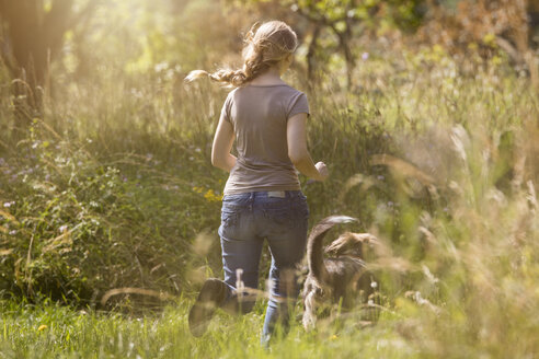 Rückenansicht einer jungen Frau, die ihrem Hund auf einer Wiese hinterherläuft - MIDF000745