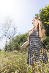 Lächelnde junge Frau steht auf einer Wiese und hört mit Smartphone und Kopfhörern Musik - MIDF000752