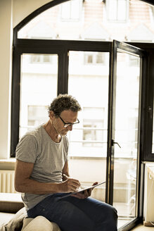 Man sitting on backrest of couch using digital tablet - FMKF002749