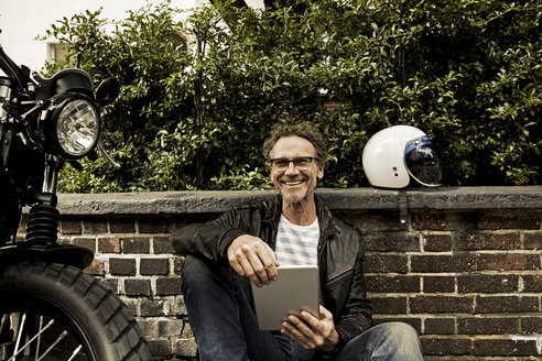 Laughing man with tablet sitting on the ground beside his motorbike - FMKF002762