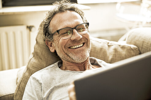 Porträt eines lachenden Mannes, der auf der Couch liegt und ein Tablet benutzt - FMKF002770