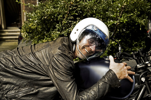 Smiling man wearing motorcycle helmet and leather jacket lying on his motorbike - FMKF002773
