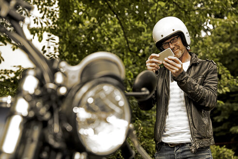 Smiling man with motorcycle helmet taking photo of his motorbike - FMKF002774