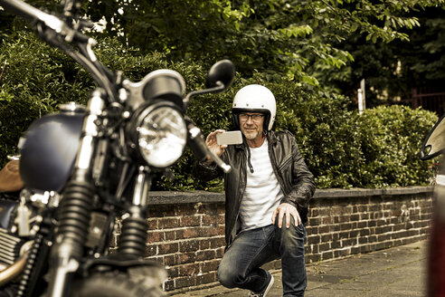 Smiling man with motorcycle helmet taking photo of his motorbike - FMKF002775