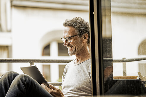 Lächelnder Mann sitzt auf einem Balkon und benutzt ein digitales Tablet - FMKF002779