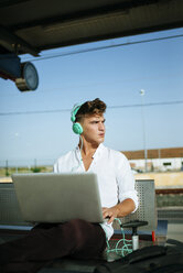 Junger Mann mit Kopfhörern und einem Laptop am Bahnsteig - KIJF000559