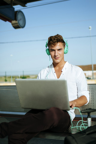 Junger Mann mit Kopfhörern und einem Laptop am Bahnsteig, lizenzfreies Stockfoto