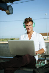Young man wearing headphones and using a laptop at station platform - KIJF000560