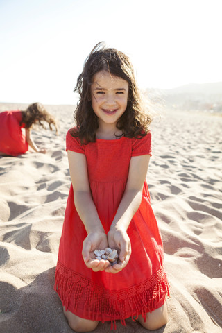 Porträt eines lächelnden kleinen Mädchens, das am Strand kniet und Muscheln in den Händen hält, lizenzfreies Stockfoto