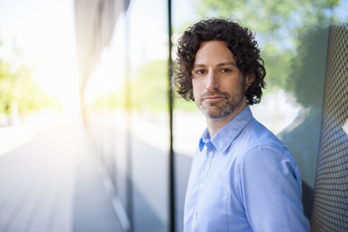 Portrait of pensive man with curly brown hair leaning against glass facade - DIGF000564