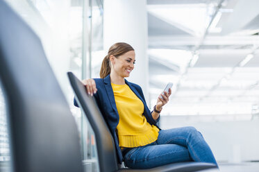 Smiling woman sitting on chair looking at cell phone - DIGF000612