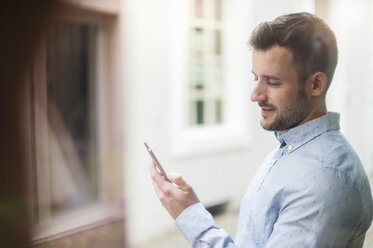 Junger Mann schaut auf sein Handy hinter einer Fensterscheibe - DIGF000683