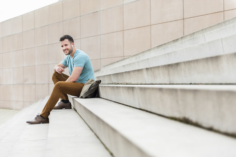 Lächelnder junger Mann im Freien, der auf einer Treppe sitzt und Musik hört, lizenzfreies Stockfoto