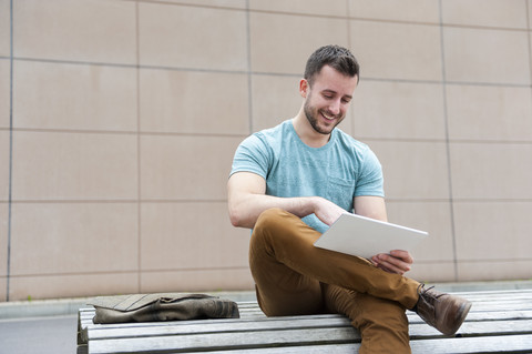 Smiling young man outdoors sitting on bench using digital tablet stock photo