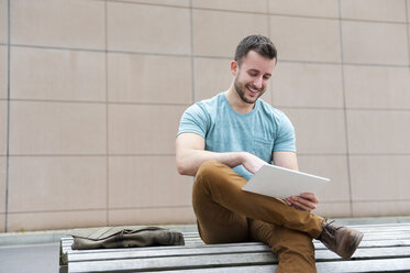 Lächelnder junger Mann im Freien, der auf einer Bank sitzt und ein digitales Tablet benutzt - DIGF000702