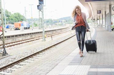 Junge Frau mit Gepäck auf Rädern schaut auf ihr Smartphone, während sie am Bahnsteig geht - DIGF000711