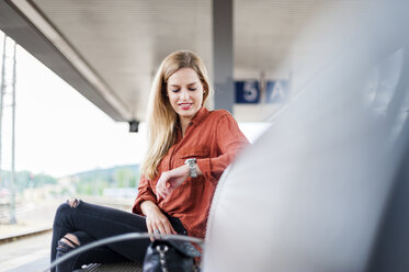 Lächelnde junge Frau, die auf einer Bank am Bahnsteig sitzt und die Zeit überprüft - DIGF000719