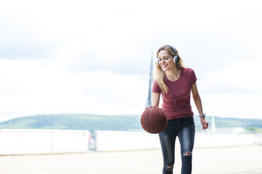 Lächelnde junge Frau mit Kopfhörern, die auf der Dachterrasse mit einem Basketball spielt - DIGF000732