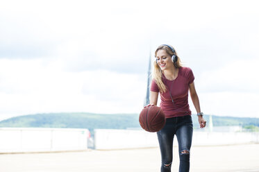 Smiling young woman with headphones playing with basketball on roof terrace - DIGF000732