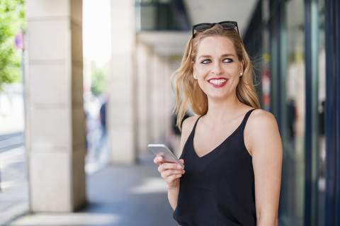 Porträt einer lächelnden jungen Frau mit Smartphone, die etwas beobachtet, lizenzfreies Stockfoto