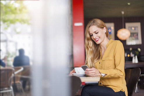 Lächelnde junge Frau, die in einem Café sitzt und auf ihr Smartphone schaut - DIGF000764