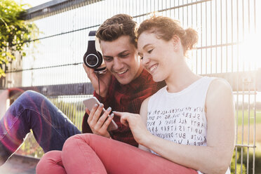 Smiling young couple at a fence with headphones and smartphone - UUF008112