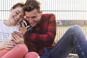 Lächelndes junges Paar an einem Zaun mit Kopfhörern und Smartphone - UUF008113