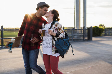 Young couple with beer bottles and skateboard kissing at sunset - UUF008131