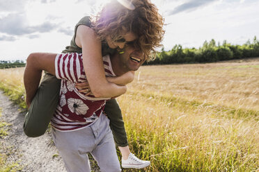Glücklicher junger Mann trägt seine Freundin huckepack auf einem Feldweg - UUF008156