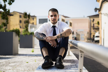 Porträt eines jungen Geschäftsmannes mit Tätowierungen auf seinen Unterarmen, der an einer Wand sitzt - GIOF001257