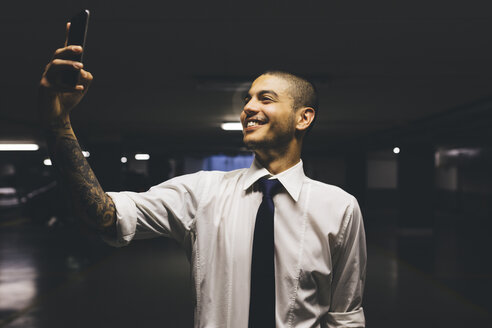 Lächelnder junger Geschäftsmann, der in einem düsteren Parkhaus steht und ein Selfie mit seinem Handy macht - GIOF001268
