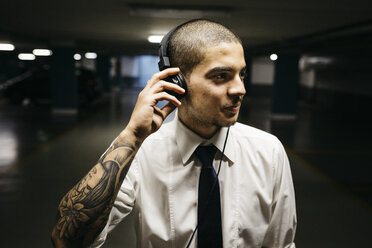 Junger Geschäftsmann mit Tätowierung auf dem Unterarm und Kopfhörern in einem düsteren Parkhaus - GIOF001270