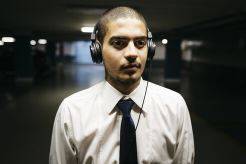Porträt eines jungen Geschäftsmannes mit Kopfhörern in einem düsteren Parkhaus - GIOF001271