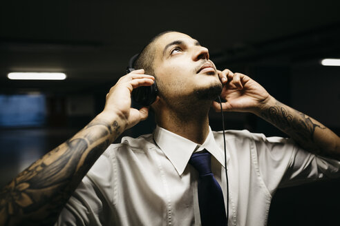 Young businessman with tattoo on his forearms with headphones in a gloomy car park - GIOF001272