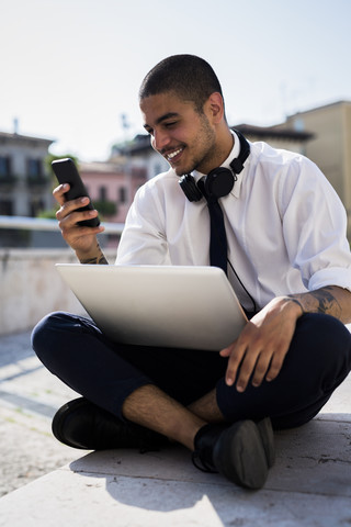 Lächelnder junger Geschäftsmann, der mit seinem Laptop an einer Wand sitzt und auf sein Handy schaut, lizenzfreies Stockfoto