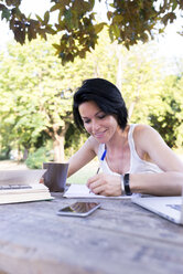 Smiling woman sitting at table in a park writing down something - FMOF000061