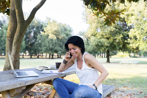Lächelnde Frau sitzt auf einer Parkbank und telefoniert mit ihrem Smartphone - FMOF000062