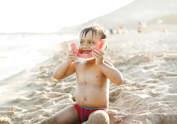 Little boy sitting on the beach at seafront eating watermelon - VABF000704