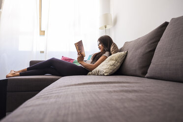 Smiling teenage girl lying on the couch reading a book - SIPF000718