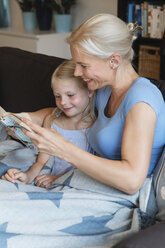 Mother and little daughter sitting on couch reading a book together - MIDF000760
