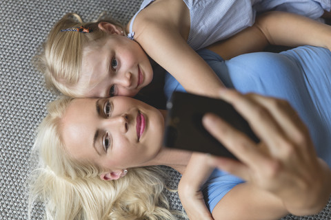 Mutter und ihre kleine Tochter liegen auf dem Boden und machen ein Selfie mit ihrem Smartphone, lizenzfreies Stockfoto