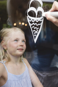 Porträt eines kleinen Mädchens hinter einer Fensterscheibe, das seine Mutter beim Bemalen einer Eistüte beobachtet - MIDF000768