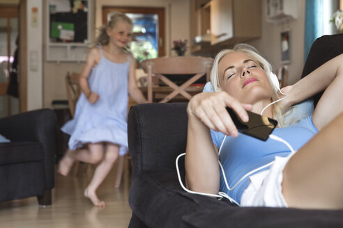 Mother lying on the couch listening music with headphones while her daughter moving in the background - MIDF000769