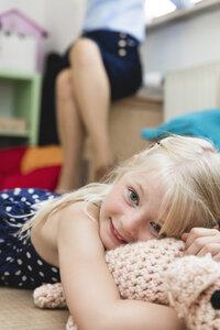 Porträt eines kleinen Mädchens mit Kuscheltier, das auf dem Boden liegt, während seine Mutter im Hintergrund sitzt - MIDF000777