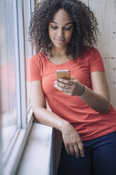 Junge Frau steht am Fenster und benutzt ein Smartphone - RIBF000470
