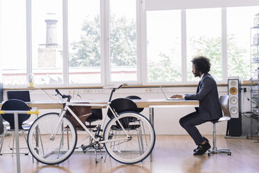 Junger Geschäftsmann bei der Arbeit im Büro mit einem an den Schreibtisch gelehnten Fahrrad - RIBF000494