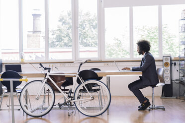 Young businessman working in office with bicycle leaning against desk - RIBF000494