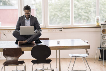 Young businessman sitting cross-legged on desk using laptop - RIBF000578